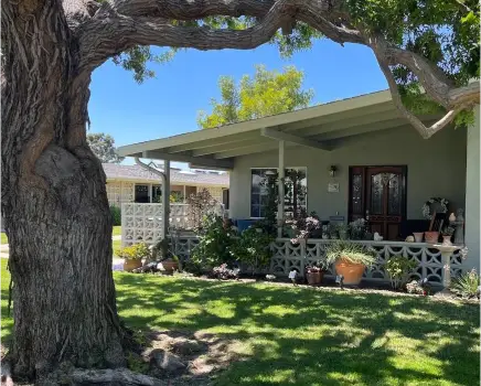 Charming exterior of a Leisure World Seal Beach home, featuring a spacious porch, lush landscaping, and a large tree, emphasizing the community's inviting atmosphere for active adults.
