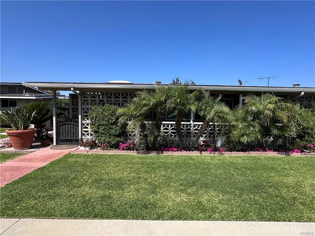 Front view of a Leisure World residence featuring a gated entry, lush landscaping with palm trees and colorful flowers, and a well-maintained lawn, highlighting the community's focus on accessibility and outdoor aesthetics.