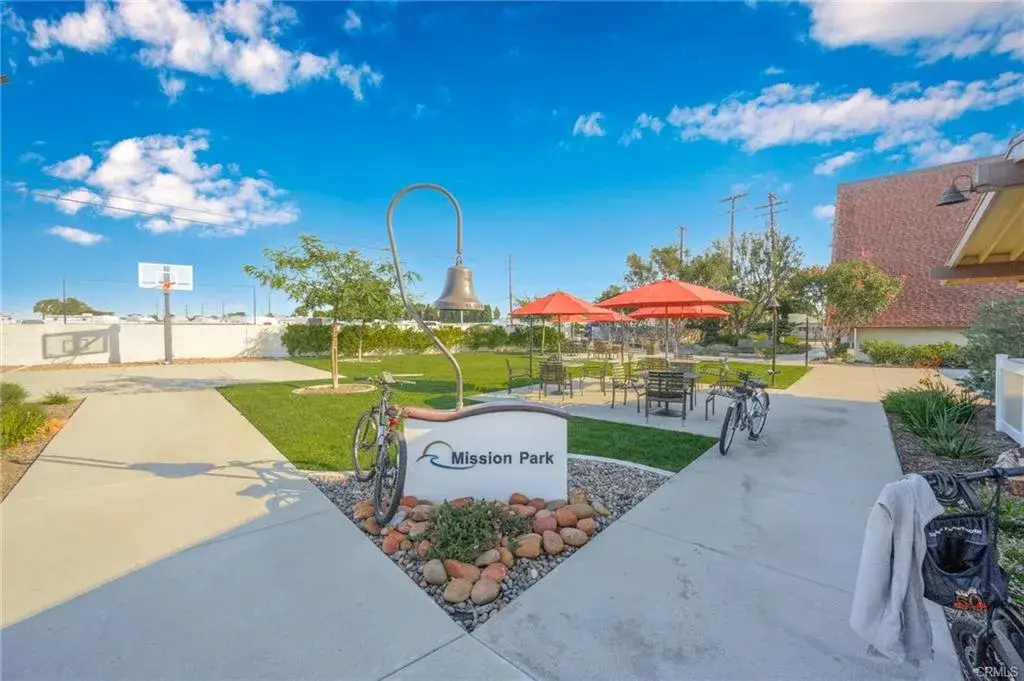 Mission Park entrance featuring a sign, outdoor seating with red umbrellas, a basketball hoop, and bicycles parked along a pathway, highlighting community amenities for an active lifestyle in Leisure World Seal Beach.