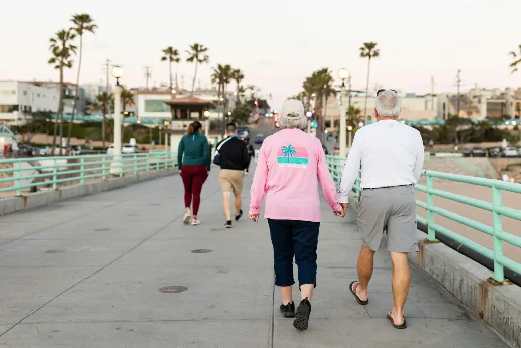 Couple walking hand in hand along a coastal promenade, enjoying the sunset, surrounded by palm trees and community atmosphere, reflecting the active lifestyle at Leisure World Seal Beach.