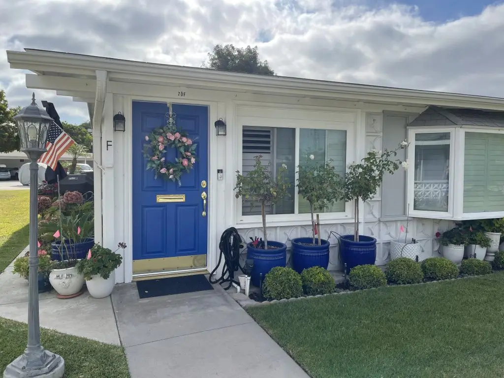 Front entrance of a Leisure World home featuring a bright blue door adorned with a floral wreath, surrounded by potted plants and a well-maintained lawn, reflecting the appealing lifestyle of the 55+ community.