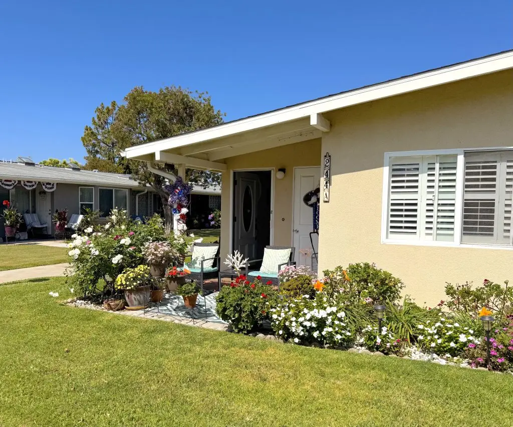 Charming Leisure World home exterior with vibrant flower gardens, inviting seating area, and clear blue sky, representing community living in a 55+ retirement community.