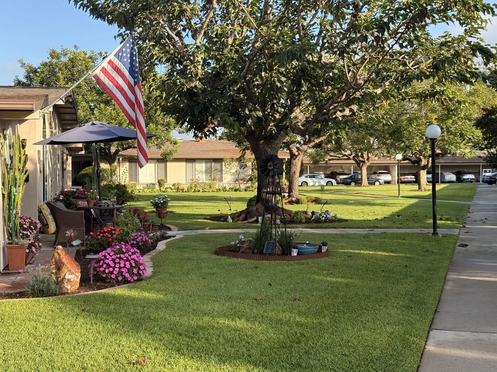 Leisure World Seal Beach community landscape featuring a well-maintained lawn, vibrant flower beds, a patio with an umbrella, and an American flag, illustrating the active adult lifestyle and outdoor amenities of the 55+ residential area.