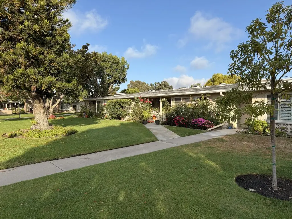 Lush green lawn with flowering shrubs and trees in front of a single-story residential building at Leisure World Seal Beach, showcasing a vibrant, welcoming community atmosphere for seniors.