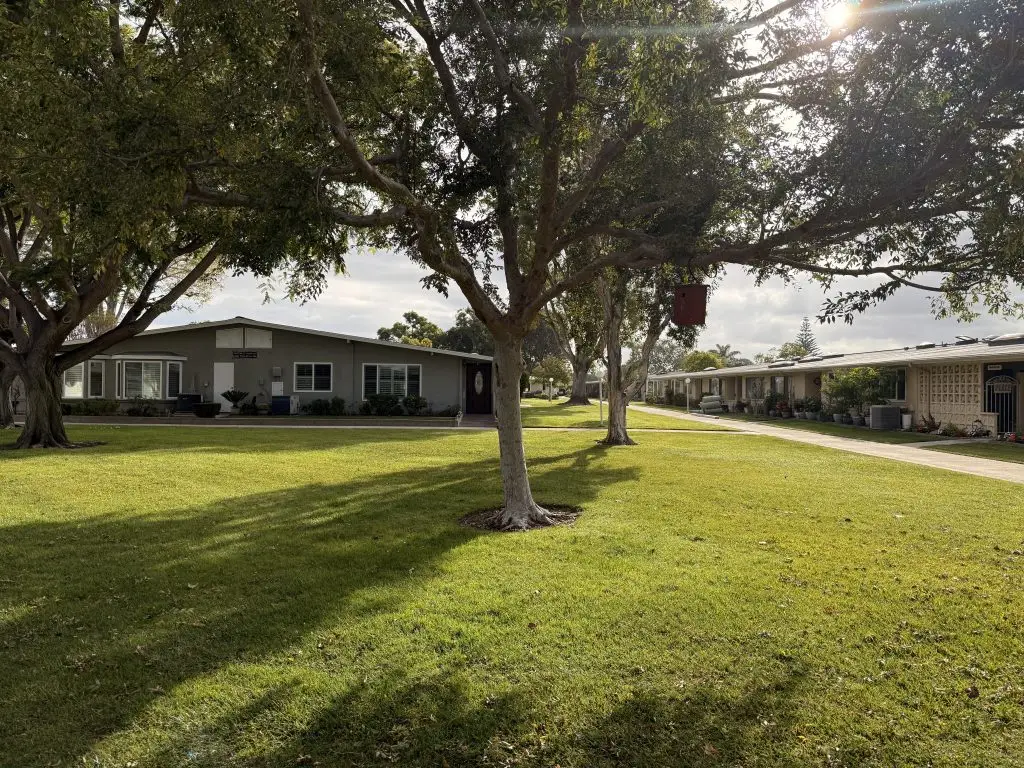 Community garden setting in Leisure World Seal Beach with well-maintained green lawn, shaded by trees, featuring residential buildings and pathways, promoting outdoor activities and social interaction for seniors.