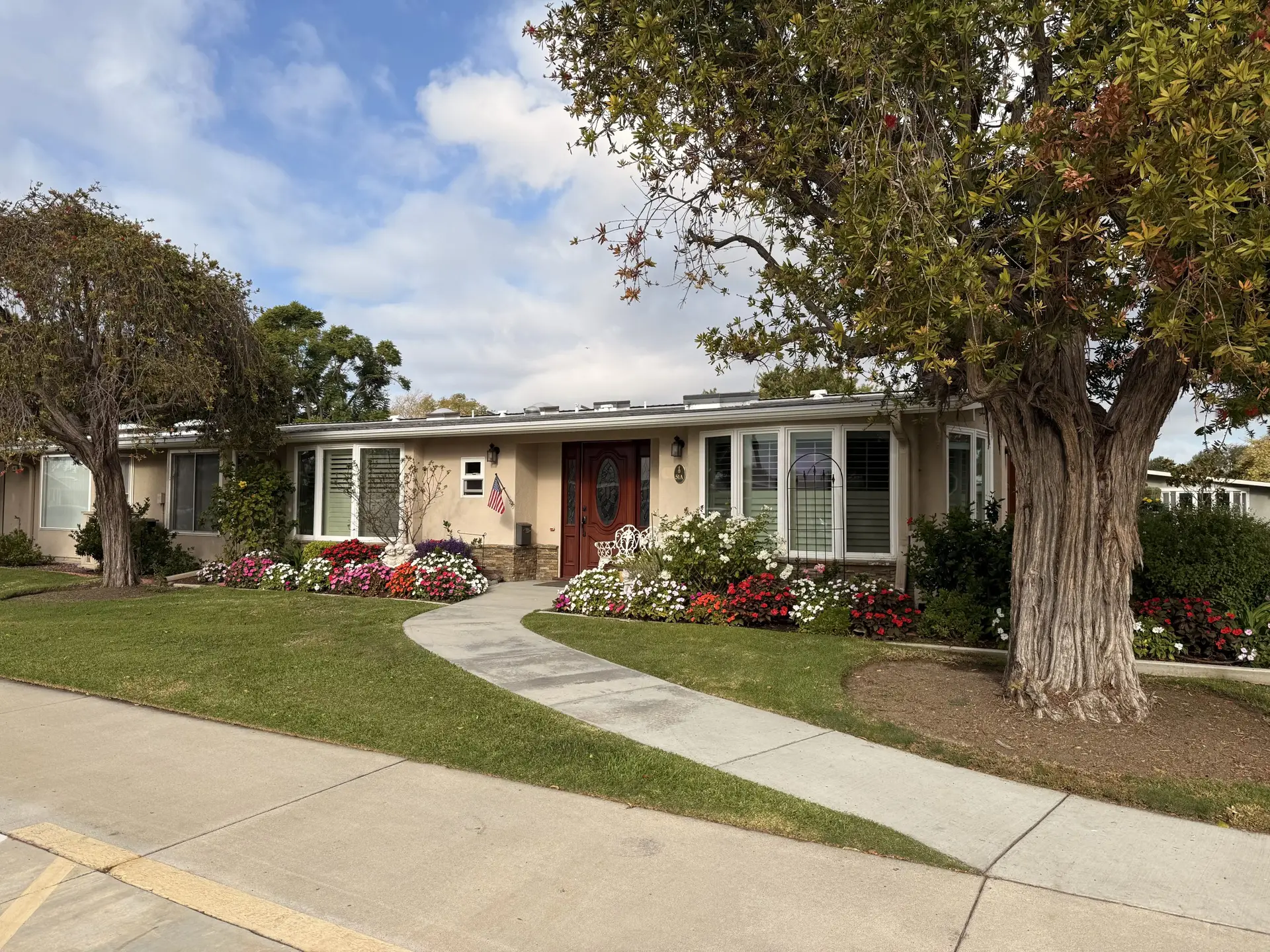 Charming single-family home in Leisure World Seal Beach, featuring a well-manicured garden with colorful flowers, a curved walkway, and a welcoming entrance with a wooden door and an American flag.