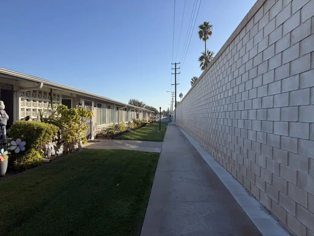 Pathway lined with well-maintained condominiums and lush landscaping at Leisure World Seal Beach, California, with a tall wall and palm trees in the background, illustrating community living and outdoor accessibility.