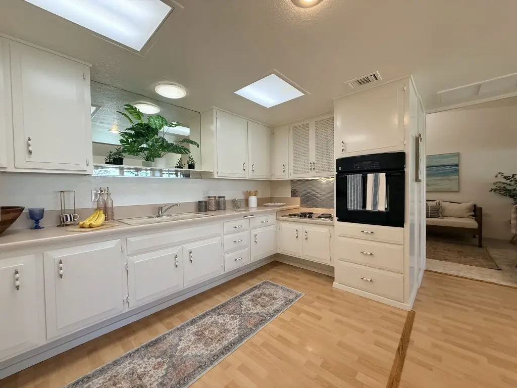 Modern kitchen with white cabinetry, stainless steel appliances, and a cozy living area visible in the background, showcasing a welcoming atmosphere ideal for Leisure World residents.